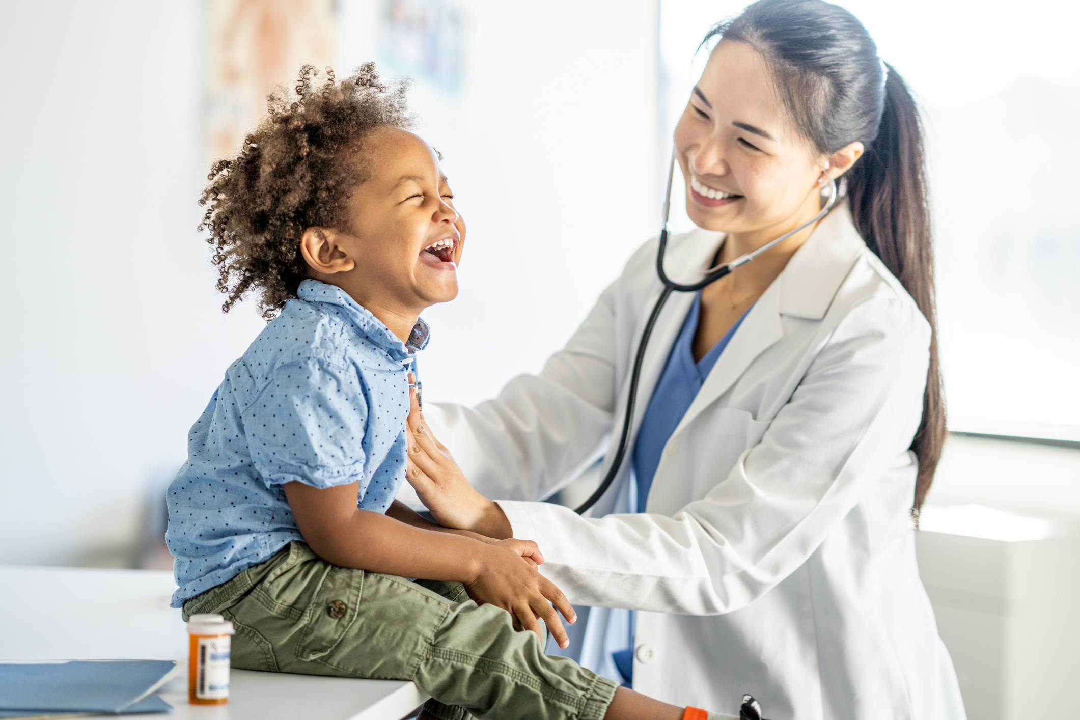 A young boy, sits up on an exam able with a big smile on his face, during a routine check-up. His doctor is leaning in with her stethoscope to listen to his heart.